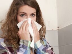 sick-woman-and-mold-on-wall-in-background a sick woman covering her nose with tissue paper and mold can be seen in the background on the wall.