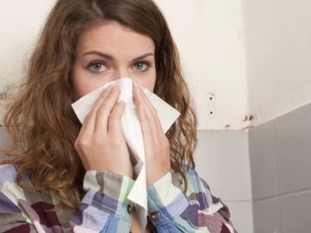 a sick woman covering her nose with tissue paper and mold can be seen in the background on the wall.